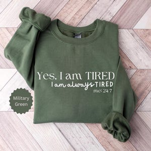 May include: Military green sweatshirt with white text that reads "Yes, I am TIRED, I am always TIRED, me: 24/7." The sweatshirt is folded on a light wood grain surface. A military green color swatch is in the lower left corner.