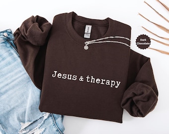 May include: Dark chocolate-colored sweatshirt with the text "Jesus & therapy" in white. A silver necklace with a pendant is draped across the front. The sweatshirt is paired with blue jeans.