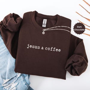 May include: Dark chocolate-colored sweatshirt with the text "jesus & coffee" in white. A silver necklace with a pendant is draped across the front. The sweatshirt is folded next to a pair of blue jeans.