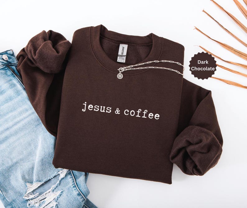 May include: Dark chocolate-colored sweatshirt with the text "jesus & coffee" in white. A silver necklace with a pendant is draped across the front. The sweatshirt is folded next to a pair of blue jeans.