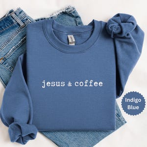 May include: Indigo blue sweatshirt with the text "jesus & coffee" in white. The sweatshirt is folded with the sleeves bunched up. A pair of blue jeans are visible in the background. A small indigo blue circle with the words "Indigo Blue" is in the bottom right corner.