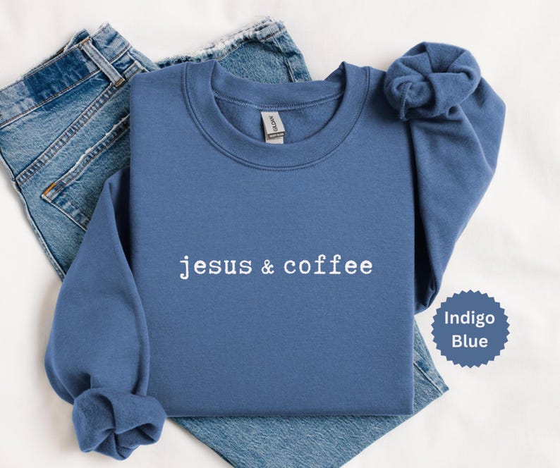 May include: Indigo blue sweatshirt with the text "jesus & coffee" in white. The sweatshirt is folded with the sleeves bunched up. A pair of blue jeans are visible in the background. A small indigo blue circle with the words "Indigo Blue" is in the bottom right corner.