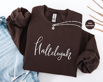 May include: A dark chocolate-colored sweatshirt with the word "Hallelujah" in white script. A silver necklace with a pendant is draped across the front. The sweatshirt is folded next to a pair of blue jeans.