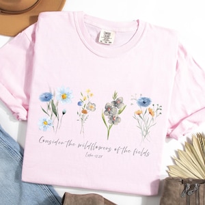 Puede incluir: Una camiseta rosa claro con un diseño floral en acuarela, con flores azules, amarillas y grises. El texto "Considerad los lirios del campo" está impreso debajo de las flores. La camiseta es de material suave.