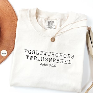 May include: An ivory short-sleeved t-shirt featuring black text that reads FGSLTWTHGHOBS TWBIHSNPBHEL and John 3:16. The shirt has a crew neckline and a Comfort Colors brand label on the inside collar. A gold-toned necklace with a small circular pendant rests on the fabric.