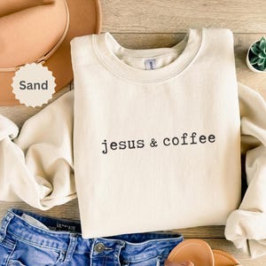 May include: Cream-colored sweatshirt with the text "jesus & coffee" in black. The sweatshirt is folded, with a tan hat and blue jeans visible. A small succulent plant in a white pot is also in the image.