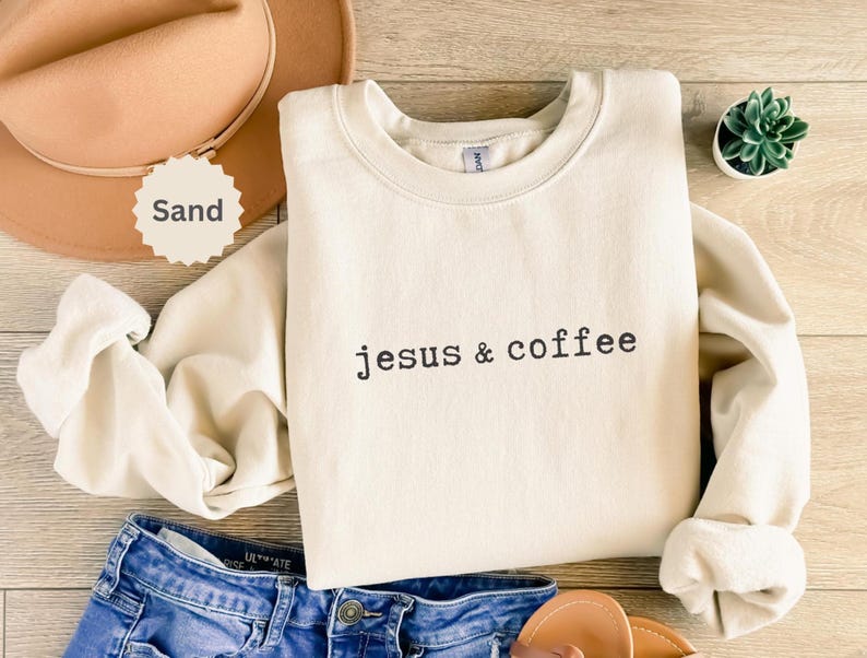 May include: Cream-colored sweatshirt with the text "jesus & coffee" in black. The sweatshirt is folded, with a tan hat and blue jeans visible. A small succulent plant in a white pot is also in the image.