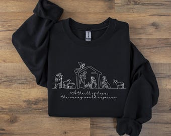 May include: Black crewneck sweatshirt featuring a white line drawing of a nativity scene. The design includes figures, a stable, and the text "A thrill of hope, the weary world rejoices" in a script font. A cozy, long-sleeved top.