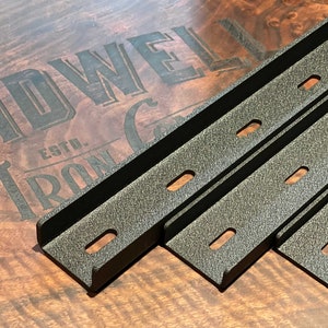 May include: Three black metal shelf brackets with a textured finish and multiple holes. The brackets are stacked on top of each other on a wooden surface. The text "Bidwell Iron Co. Est." is visible in the background.