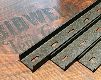 Peut inclure: Trois supports d'étagère en métal noir avec une finition texturée et plusieurs trous. Les supports sont empilés les uns sur les autres sur une surface en bois. Le texte "Bidwell Iron Co. Est." est visible en arrière-plan.