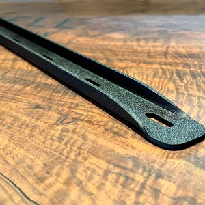 May include: A close-up shot of a black metal object with a textured surface. The object has a long, narrow shape with a curved end and several small holes. The object is set against a wooden background.
