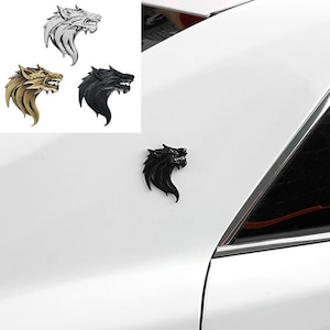 Op de afbeelding: Een zwart wolfskop-embleem bevestigd op het witte buitenpaneel van een voertuig. Het ontwerp toont een gestileerd, grommend wolfsprofiel met scherpe details. Aanvullende zilveren, gouden en zwarte versies van het embleem zijn hierboven getoond.