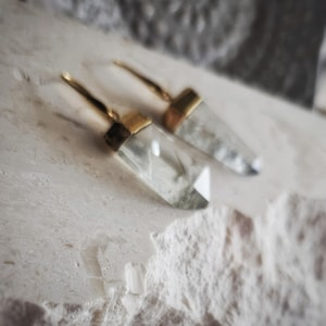 May include: A pair of clear crystal earrings with gold-colored accents. The earrings have a geometric, faceted design and are displayed on a textured, off-white surface. The earrings are a type of jewelry.
