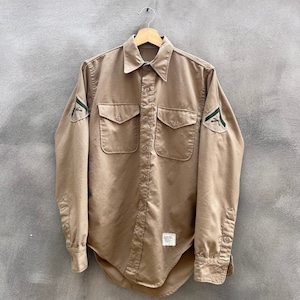 Puede incluir: Una camisa de manga larga de color beige con dos bolsillos de parche en la parte delantera y cierre de botones. La camisa tiene parches verdes y blancos en las mangas.