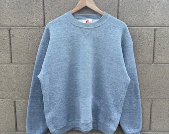 May include: A heather gray crew neck sweatshirt hanging on a wooden hanger. The sweatshirt has a classic, relaxed fit with ribbed cuffs and hem. The brand logo is visible on the inside collar. The background is a textured brick wall.