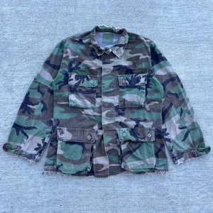 Könnte beinhalten: Jacke mit Tarnmuster mit vier Vordertaschen und zwei Brusttaschen. Die Jacke hat eine Knopfleiste, einen Kragen und lange Ärmel. Das Farbschema umfasst Grüntöne, Braun und Schwarz.