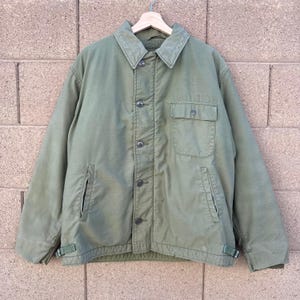May include: An olive green jacket with a button-up front, a collared neckline, and two front pockets. The jacket has a military-style design, with a small pocket on the chest and adjustable straps at the waist. The text "VA 710" is visible on the right side.
