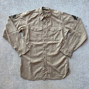 May include: A tan long-sleeved shirt with two pockets and button fastenings. The shirt has black chevron patches on each shoulder.