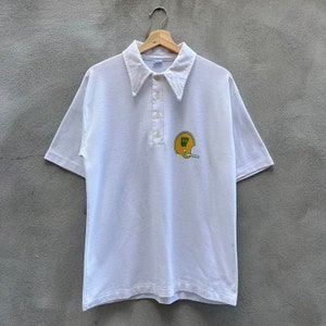 Puede incluir: Polo blanco con mangas cortas y cuello ancho. La camisa presenta un pequeño emblema de un casco de fútbol americano amarillo con detalles verdes. La camisa está colgada en una percha de madera sobre un fondo gris.