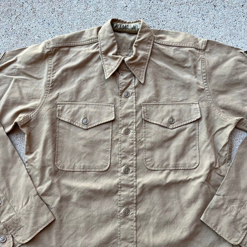 May include: A tan long-sleeved button-down shirt with two large flap pockets on the front. The shirt has a classic collar and a button closure.