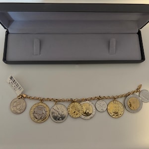 May include: A gold-toned charm bracelet with various coin-shaped charms. The bracelet is displayed near an open, black jewelry box with a gray interior. A price tag is attached to the bracelet.