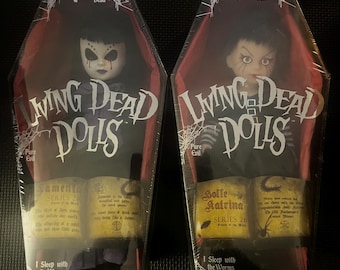 Puede incluir: Dos muñecas Living Dead Dolls en un embalaje con forma de ataúd. Las muñecas están vestidas de negro y tienen la piel pálida, el pelo negro y un maquillaje oscuro. Las muñecas sostienen libros con texto que dice "Samenta Series 20" y "Bolle Katrina Series 20". El embalaje tiene el texto "Living Dead Dolls" y "I Sleep with the Worms".