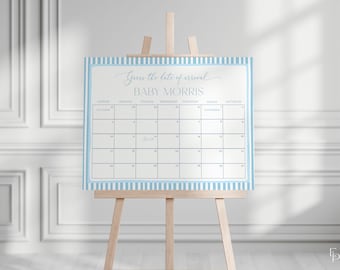 Op de afbeelding: Een lichtblauw en wit gestreepte babyshowerkalender op een houten ezel. De kalender heeft een lichtblauwe rand en de tekst "Guess the date of arrival" en "BABY MORRIS" in een scriptlettertype. De kalender is voor de maand oktober.