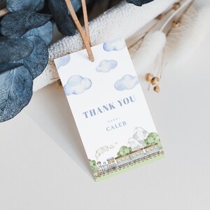 Puede incluir: Una etiqueta blanca con nubes azules de acuarela y una ilustración de tren. La etiqueta dice "THANK YOU" y "love, CALEB" en texto azul. Una cuerda de cuero marrón está adjunta.