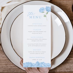 May include: A blue and white menu card with a floral design. The menu card lists the appetizers, starter, main course, dessert, and the date of the event. The text on the menu card reads "The Menu", "Appetizer", "Starter", "Main Course", "Dessert", "Charlotte's Bridal Shower", and "April 18, 201X".