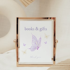 Puede incluir: Un cartel con marco dorado con el texto "libros y regalos" y una ilustración de mariposa morada. El cartel también incluye el texto "gracias" en cursiva.