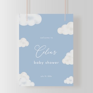 May include: A baby shower sign with a light blue background and white cloud illustrations. The sign reads "welcome to Celia's baby shower" with the date July 15, 202x. The sign is hanging from a brown string.
