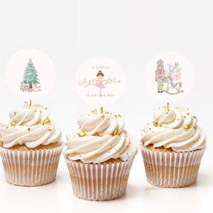 May include: Three cupcakes with white frosting and gold sprinkles, each topped with a decorative round image. The images feature a Christmas tree, a ballerina, and a nutcracker with a rocking horse. The text on the middle image says "A Little Sugar Plum Is On The Way."
