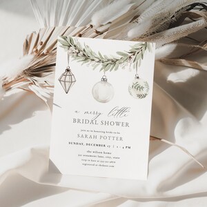 May include: A white invitation card for a bridal shower with the text "a merry little BRIDAL SHOWER". The card features watercolor illustrations of hanging ornaments and a garland. The card is on a white fabric background.