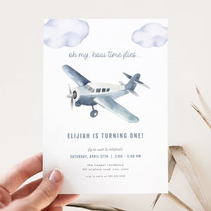 May include: A blue and white airplane illustration with clouds on a white background. The text "oh my, how time flies..." is at the top of the invitation. The text "ELIJAH IS TURNING ONE!" is in the middle of the invitation. The text "fly on over to celebrate" is below the airplane. The text "SATURDAY, APRIL 27TH | 2:00 - 5:00 PM" is below the text "fly on over to celebrate". The text "the cooper residence" is below the date and time. The text "321 airplane road, city, state" is below the residence name.