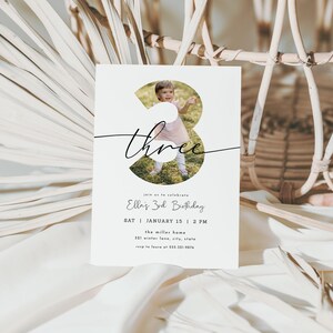 May include: A white invitation card with a photo of a child inside the number three. The word "three" is written in cursive. The text below reads "Ella's 3rd Birthday" with the date, time, and location.