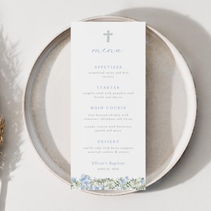 May include: A white menu card with a cross at the top, listing a meal for a baptism. The menu is on a gray plate, with a dried plant on the left. The menu includes appetizers, starters, main course, and dessert.
