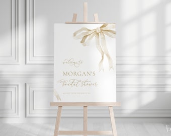May include: A white sign with a watercolour bow in gold tones. The sign reads "Welcome to Morgan's Bridal Shower 42 Days Until She Says I Do".