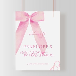 May include: A white sign with a pink watercolor bow at the top. The sign says "Welcome to Penelope's Bridal Shower 41 Days Until She Says I Do".