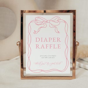 May include: A gold-framed sign with a pink ribbon design and the text "Diaper Raffle" in pink lettering. The sign also includes the text "Please place your diapers and raffle cards here. Thank you & good luck!"