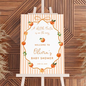 May include: A baby shower welcome sign on an easel. The sign features peach-colored stripes and a peach-themed design. Text reads "A Little Cutie is on the way" and "Welcome to Olivia's Baby Shower, June 25, 202X."