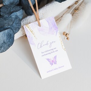 Puede incluir: Una etiqueta de agradecimiento morada y dorada con un diseño de mariposa. La etiqueta dice "Gracias por celebrar a nuestro pequeño encantador, la baby shower de Andrea."