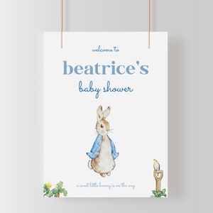 May include: A white baby shower sign with blue text that reads "welcome to beatrice's baby shower" and a watercolor illustration of Peter Rabbit in a blue jacket. The text at the bottom reads "a sweet little bunny is on the way".