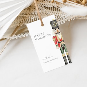 May include: White gift tag with a watercolor illustration of a nutcracker. The text reads "Happy Holidays" and "with love, The Evans Family".