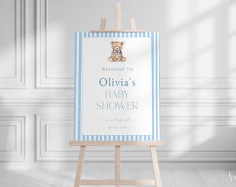 May include: A baby shower welcome sign on a wooden easel. The sign has a blue and white striped border and features a watercolor teddy bear with a blue bow tie. Text reads "Welcome to Olivia's Baby Shower" and the date.