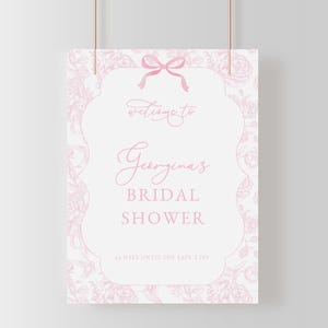 May include: A pink and white bridal shower sign with a floral design. The sign says "Welcome to Georgina's Bridal Shower" in elegant script. A pink bow is at the top, and the bottom reads "14 days until she says 'I do'."