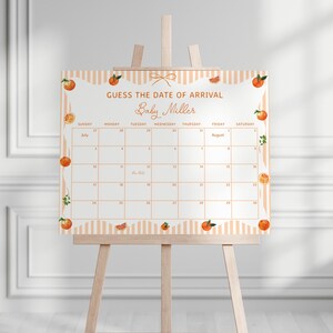 May include: A baby shower calendar on a wooden easel. The calendar is decorated with orange fruit illustrations and the text "Guess the Date of Arrival Baby Miller". The calendar has a grid for the months of July and August.