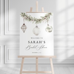 May include: A white welcome sign with a green leafy garland and hanging ornaments. The sign reads "Welcome to Sarah's Bridal Shower, December 1st, 2023".