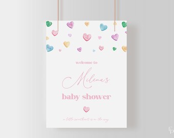 May include: A white baby shower sign with pink text that reads "welcome to Milena's baby shower a little sweetheart is on the way". The sign is decorated with colorful candy hearts.