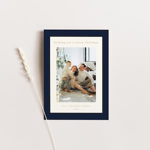 Puede incluir: Una tarjeta de Navidad con un borde azul marino que presenta una foto de una familia de tres. La familia est&aacute; sentada en una alfombra blanca en una habitaci&oacute;n con una pared blanca y una ventana. El texto "Wishing you a merry Christmas" est&aacute; impreso en dorado en la parte superior de la tarjeta. El texto "Love, the Parker family 2022" est&aacute; impreso en dorado en la parte inferior de la tarjeta.