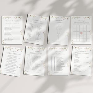 May include: A collection of eight white paper cards with floral border designs. The cards feature bridal shower game titles such as "Who Knows the Bride Best" and "Bridal Bingo". The cards are arranged on a white surface.